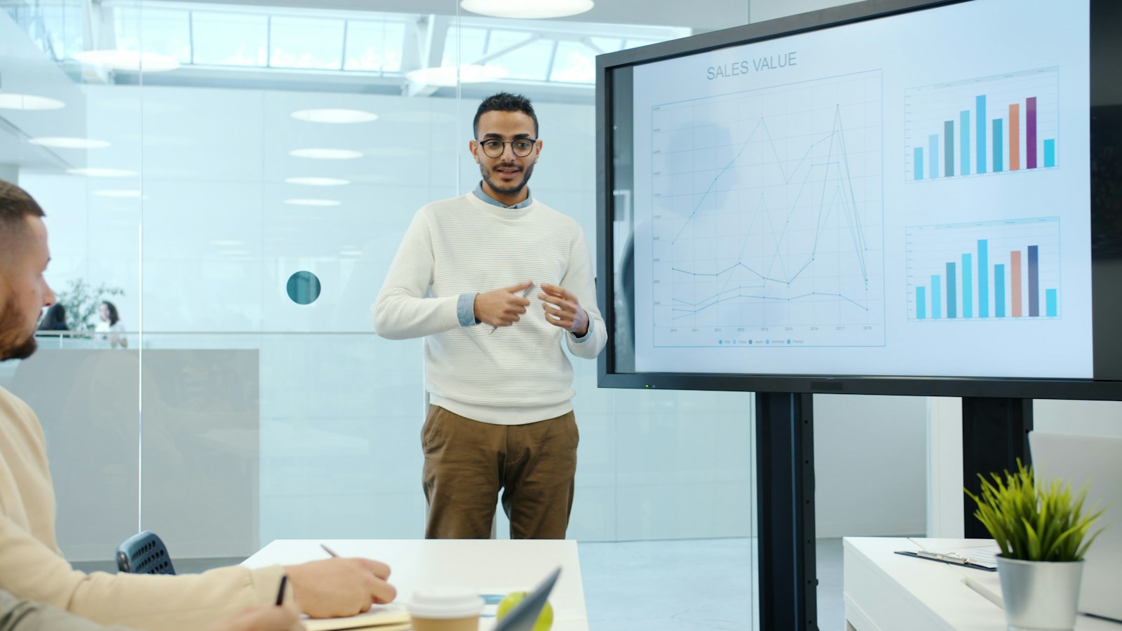 A man in a white sweater presents line and bar charts on a large screen to a colleague taking notes in a glass-walled office