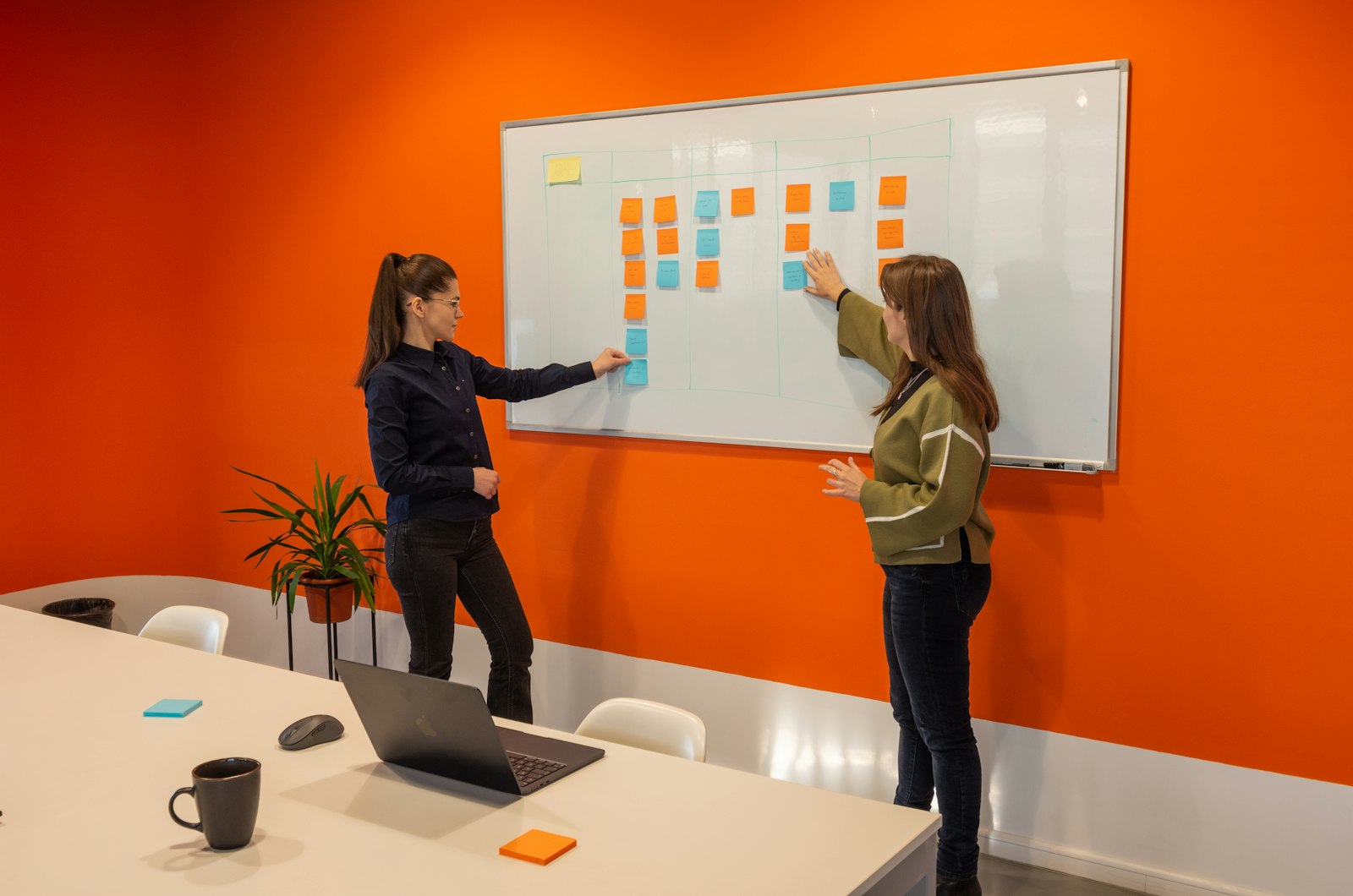 Two women arranging orange and blue sticky notes in columns on a whiteboard in an orange meeting room with a laptop on the table