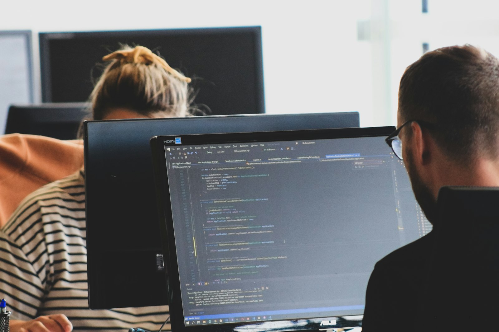Two developers at desks in a bright office, one looking at code on a monitor