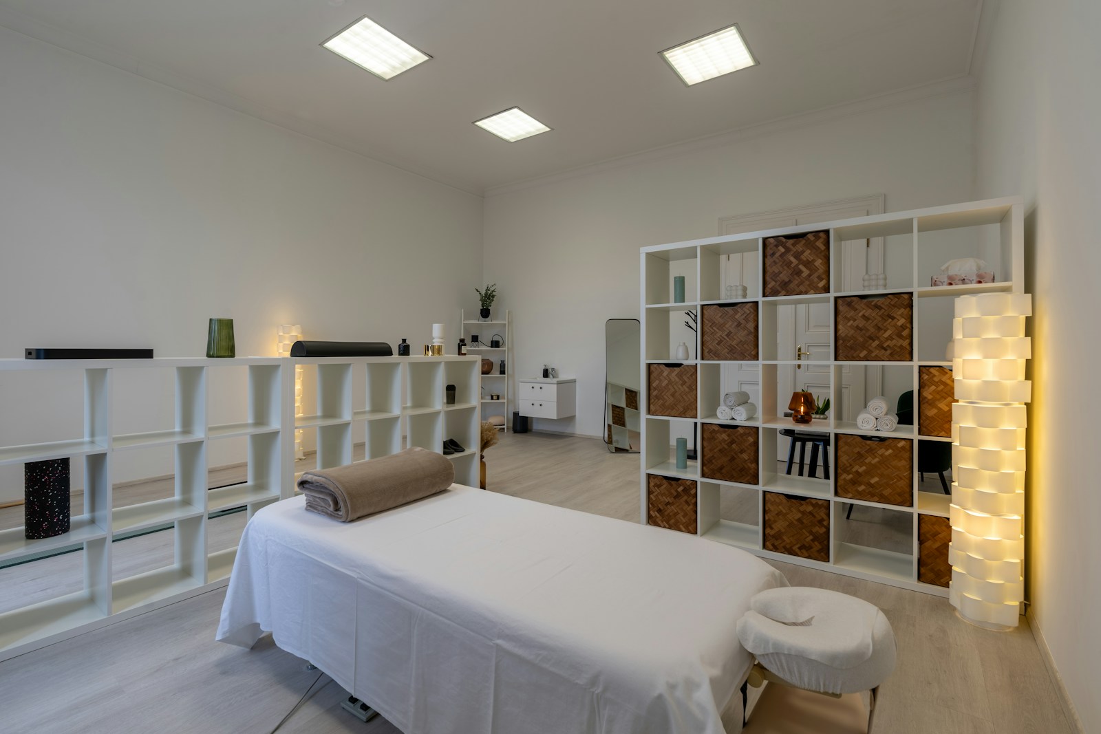 A bright massage room with a white treatment table, a folded blanket, rolled towels on white shelves and a glowing floor lamp