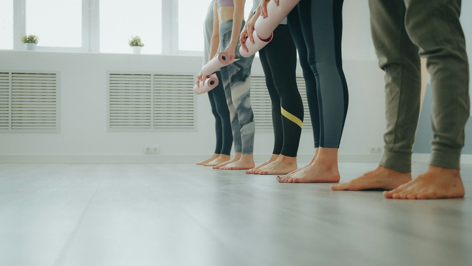 A row of barefoot people in leggings holding rolled pink yoga mats, standing on a pale floor in a bright studio