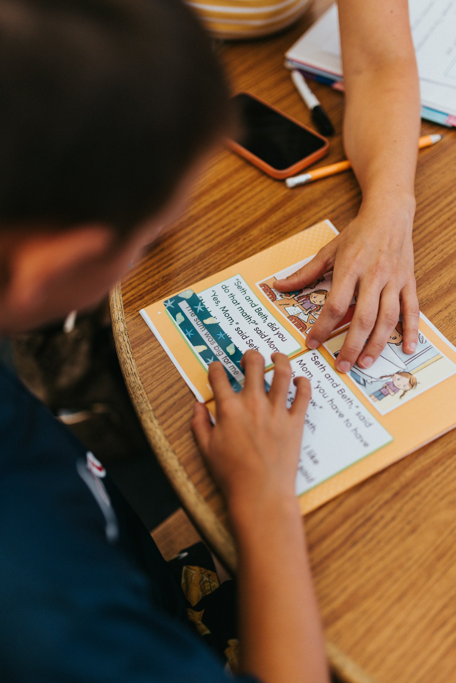 A child's hand and an adult's hand rest on an early reader booklet with cartoon pictures on a round wooden table