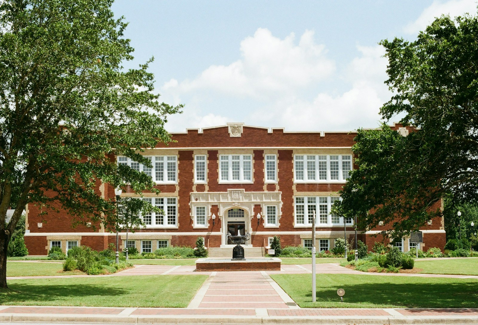 A two-story red brick school building with white trim, a fountain and a brick walkway between green lawns and trees
