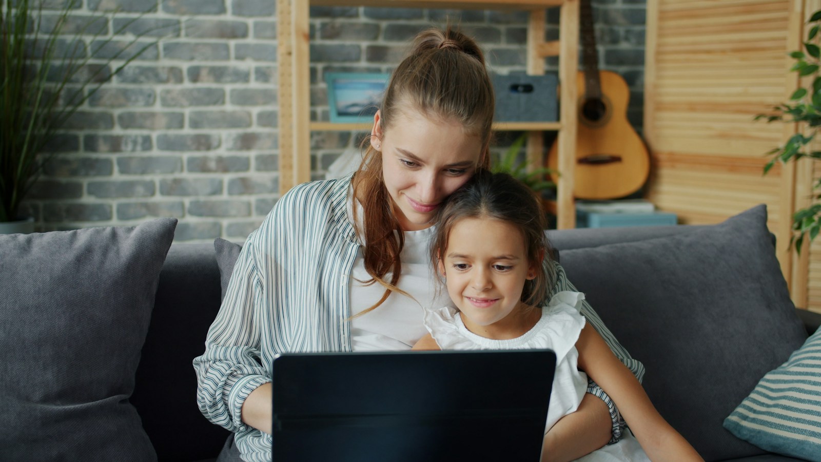A mother and her young daughter smile at a laptop together on a grey sofa at home