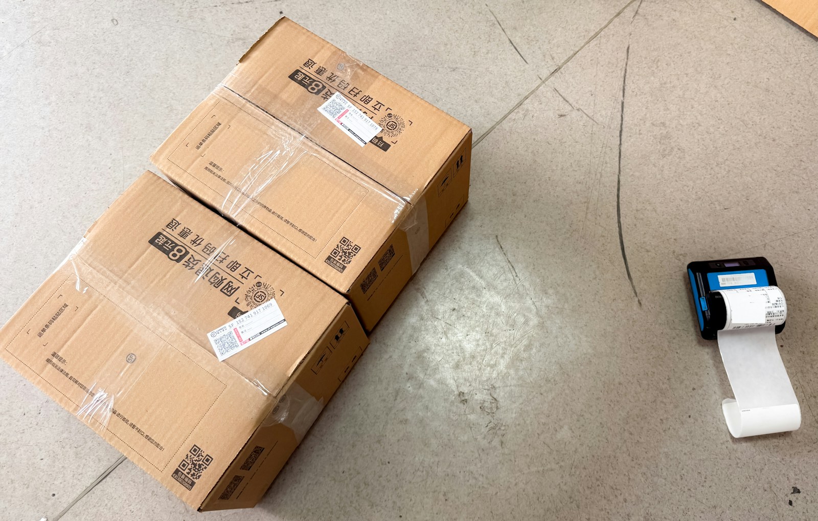 Two taped brown cardboard shipping boxes on a warehouse floor next to a portable thermal label printer