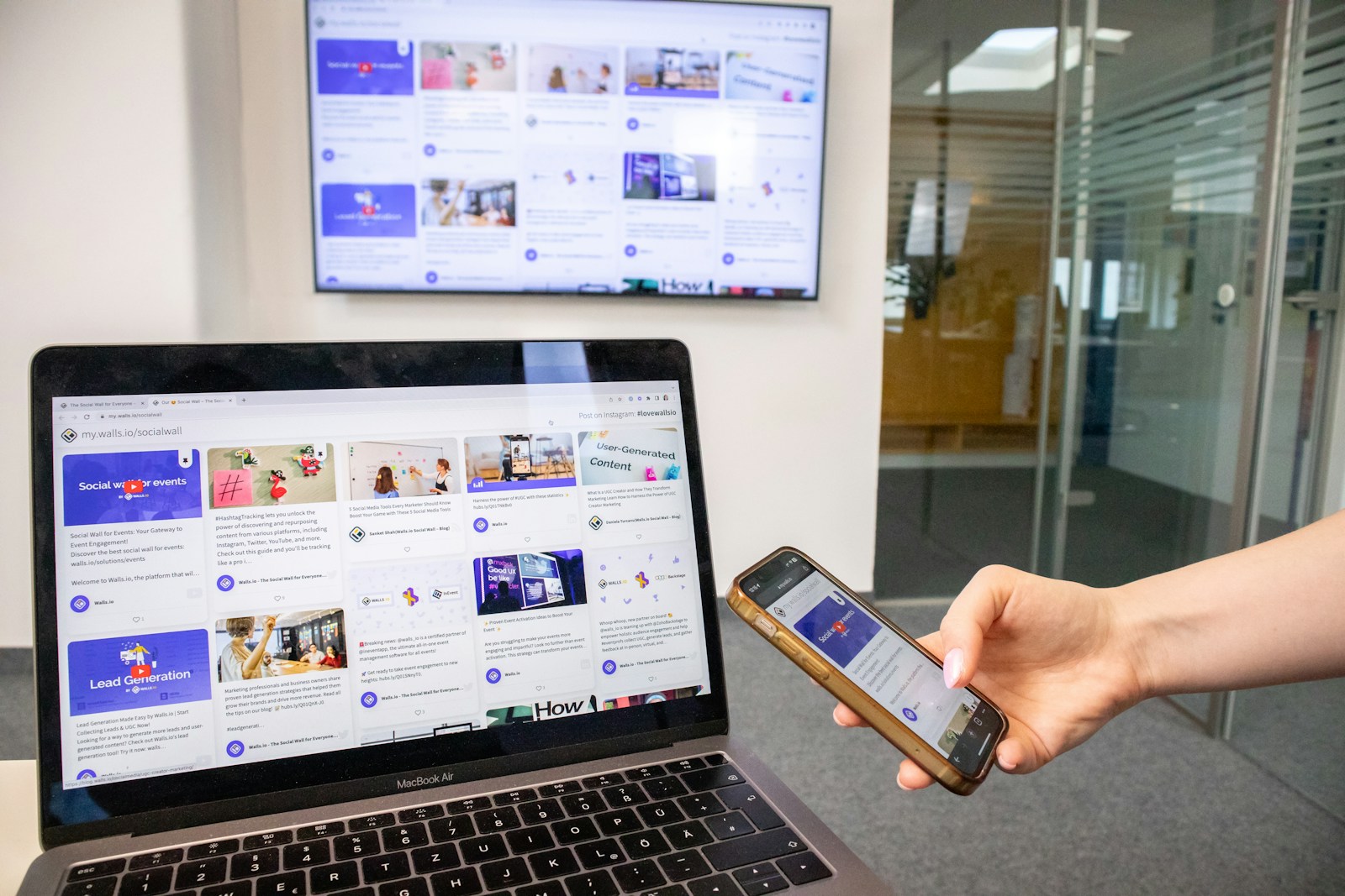 A hand holding a smartphone in front of a laptop that displays a website, in a bright office
