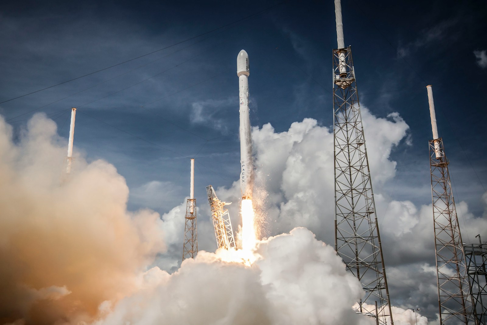 A SpaceX rocket lifting off through white smoke between launch towers under a dark blue sky