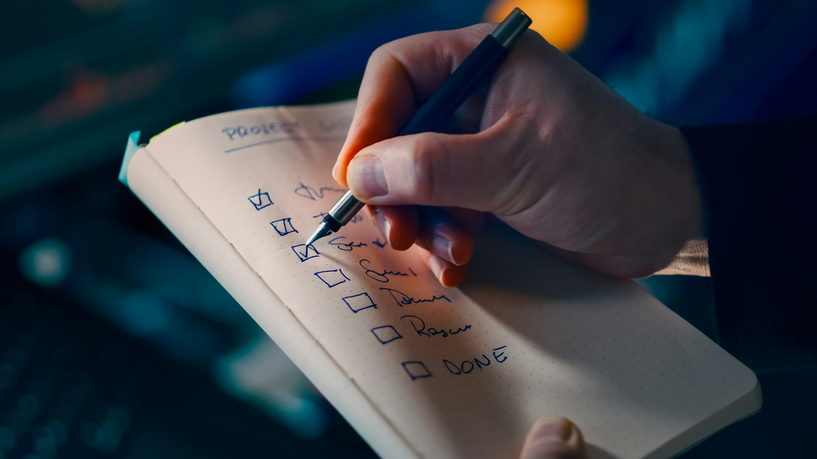 A hand with a fountain pen ticking boxes on a handwritten checklist in a small notebook