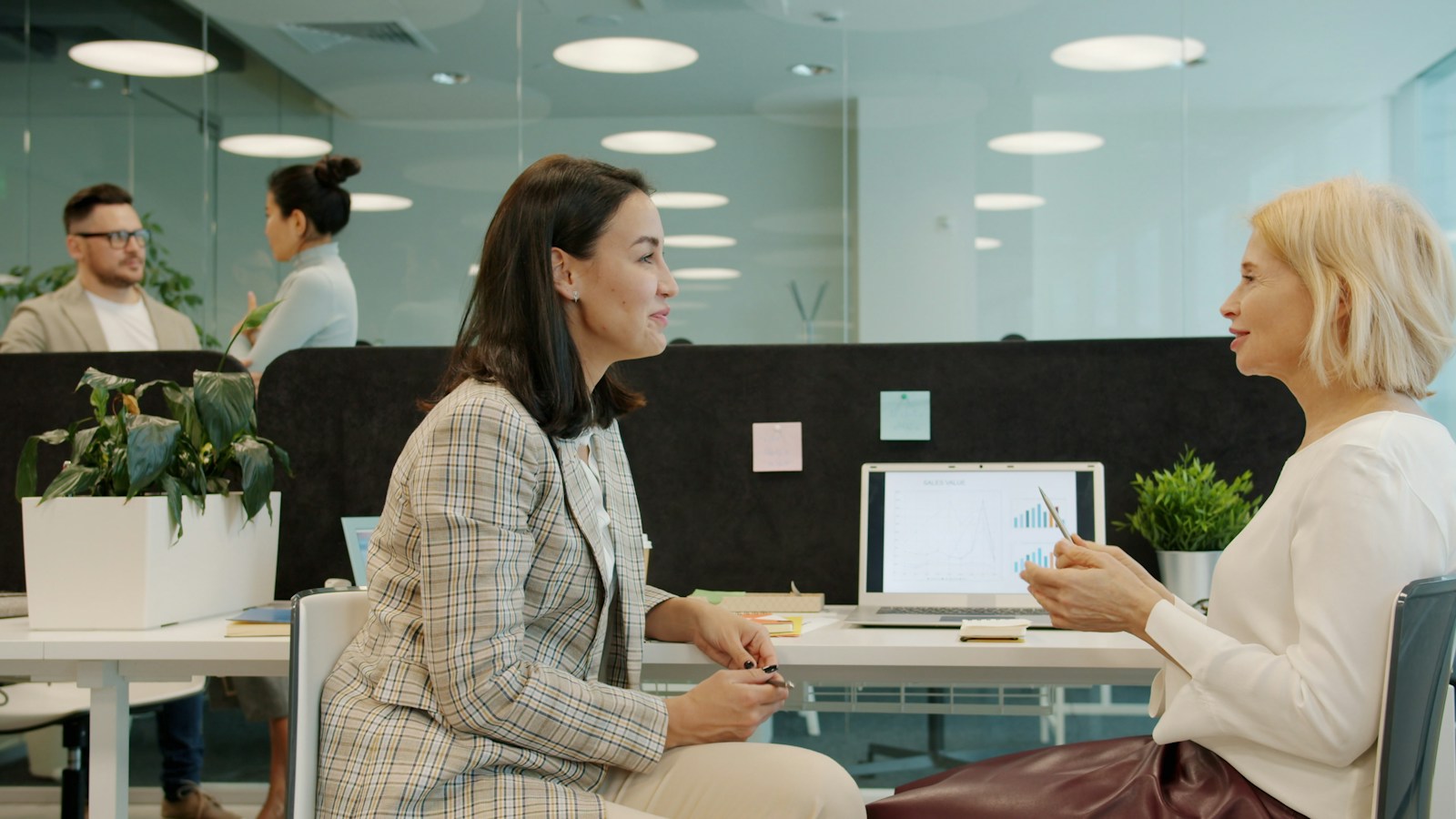 Two women in a bright open-plan office talk across a desk with a laptop showing sales charts between them