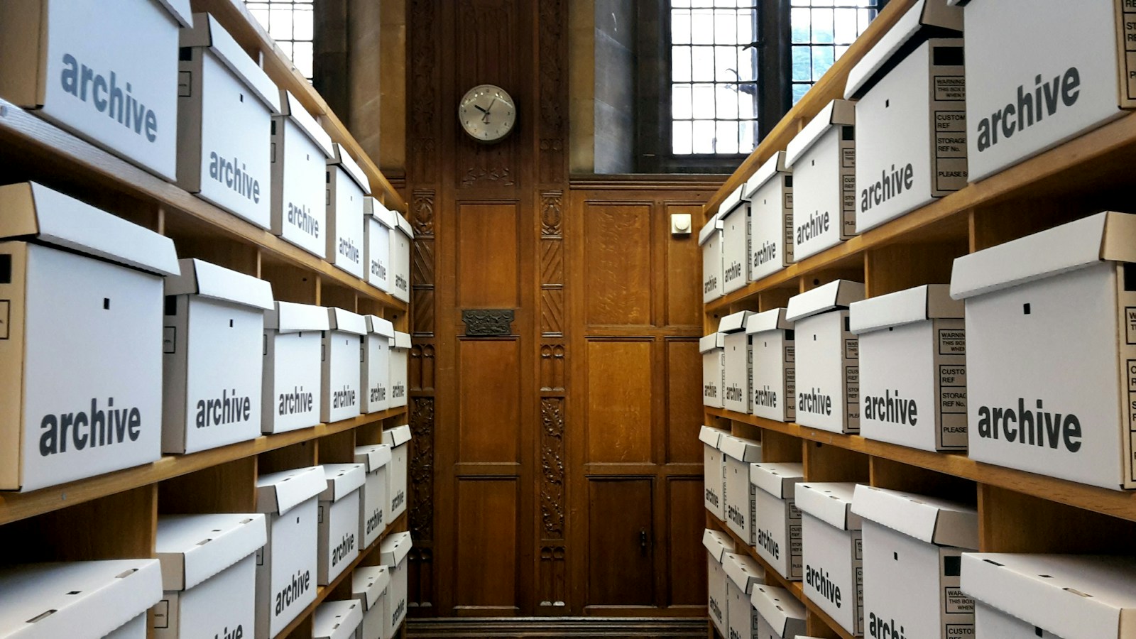 Rows of white storage boxes labelled archive fill wooden shelves on both sides of a narrow wood-panelled room