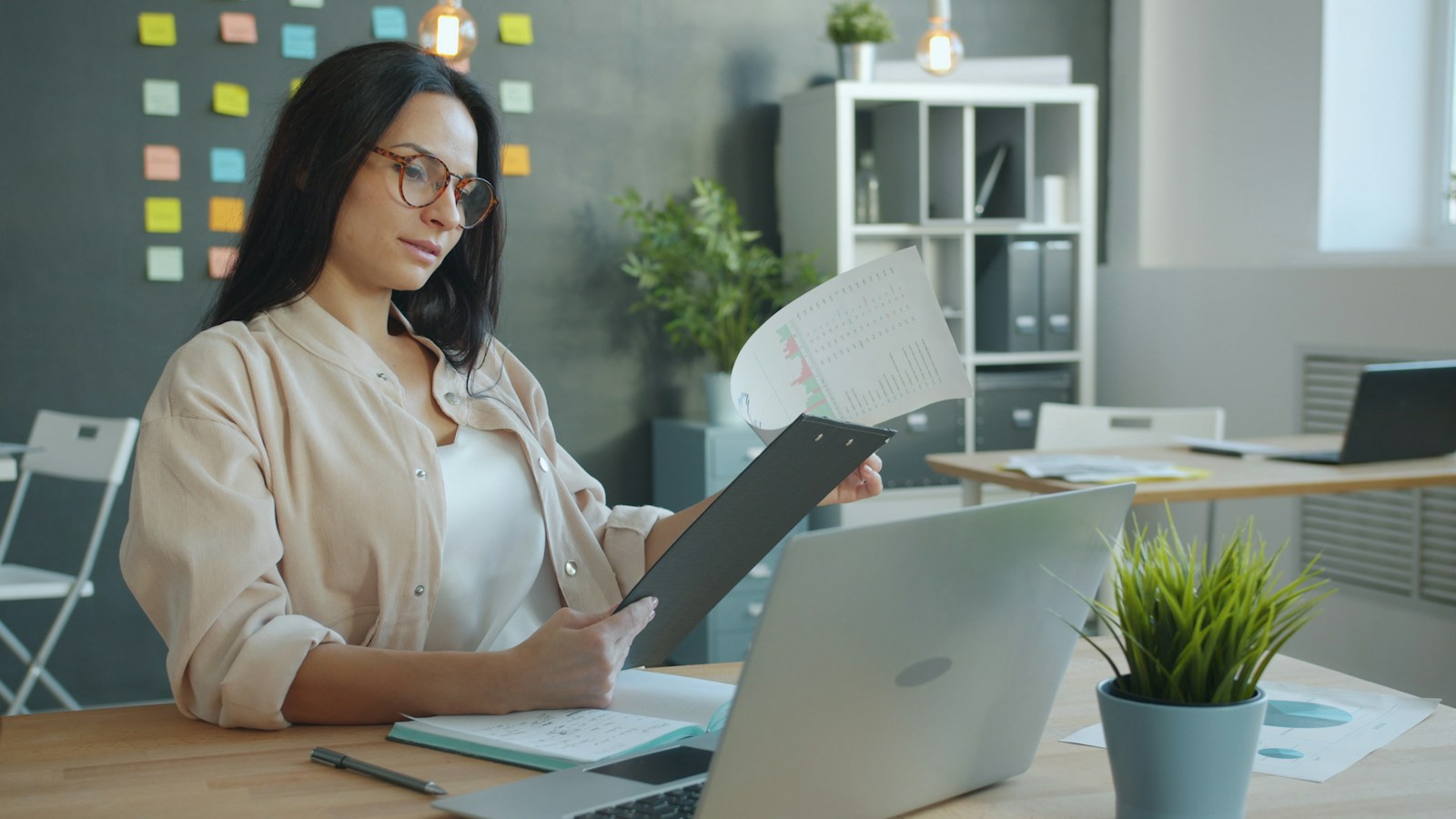 A woman in glasses reads a printed chart on a clipboard at a desk with a laptop and a notebook