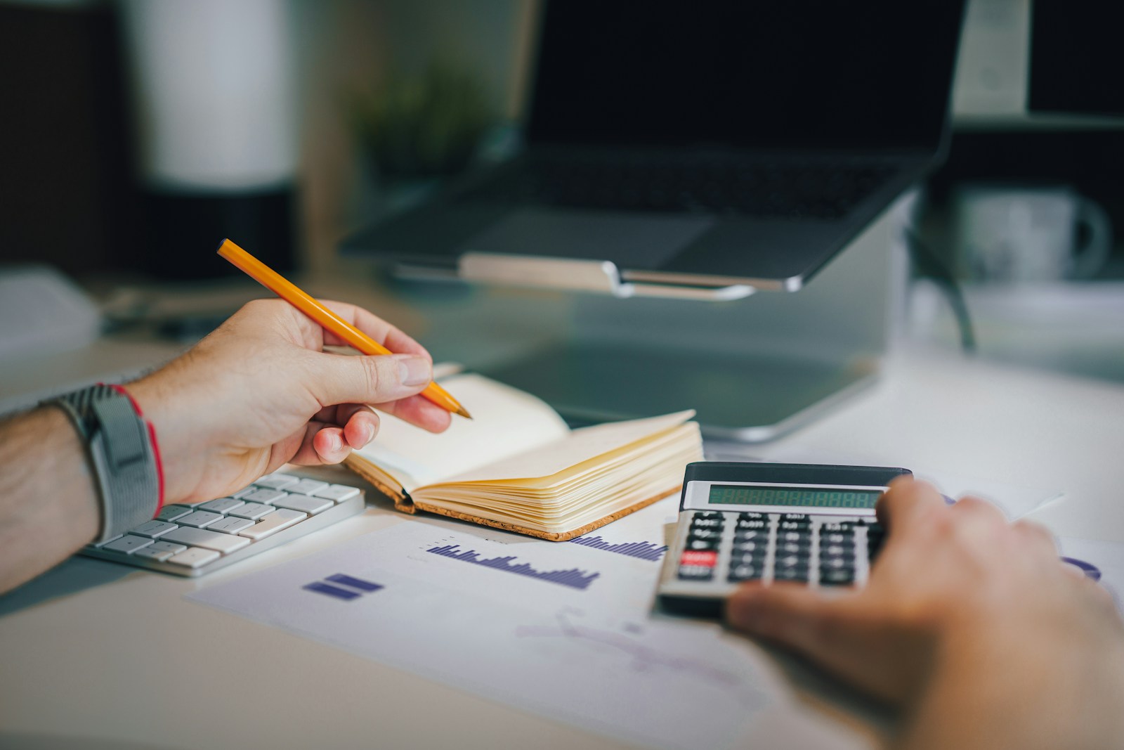 A hand holding a pencil over an open notebook beside a calculator, a printed bar chart and a laptop