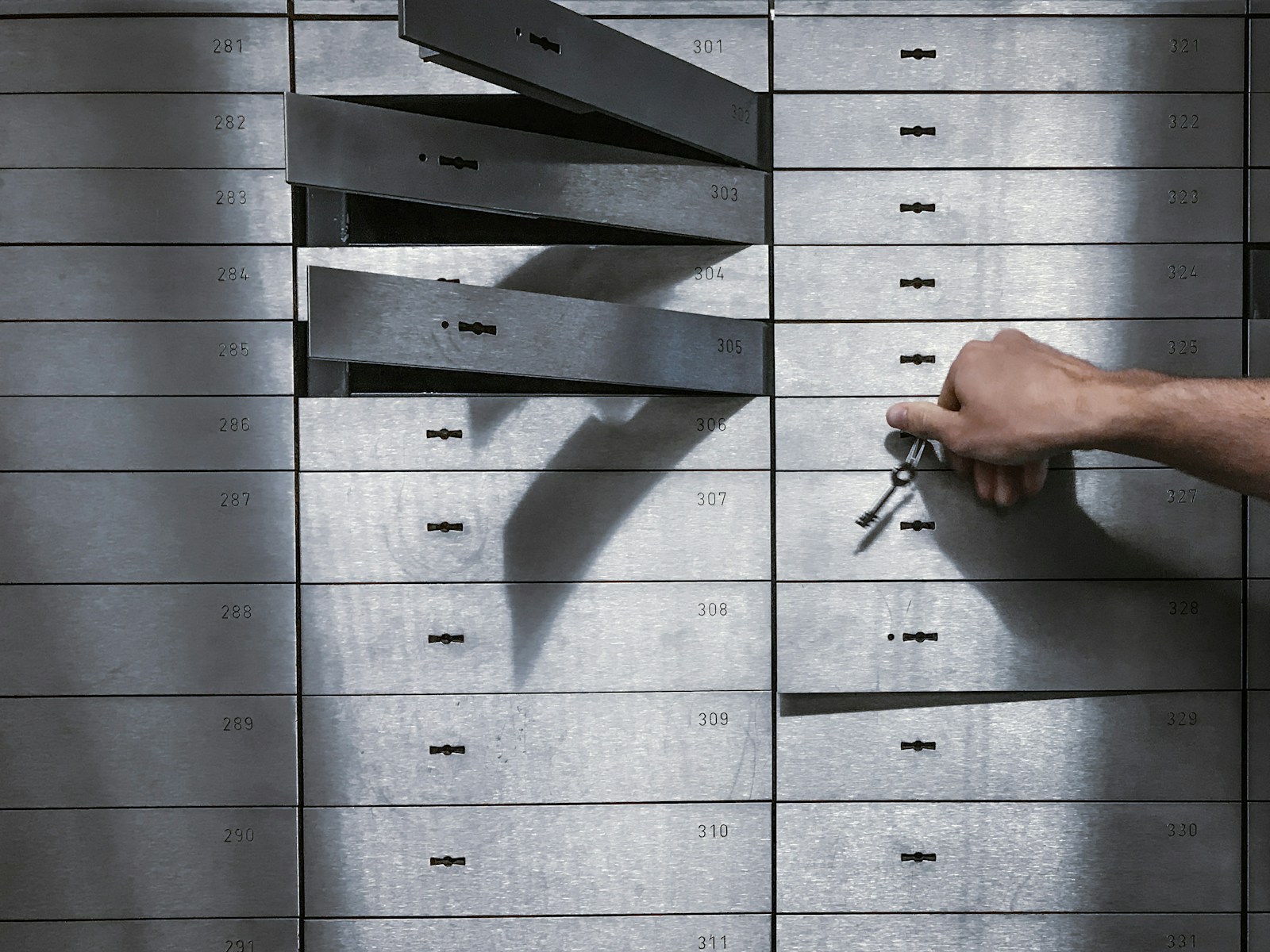 A hand turning a small key in one numbered safe deposit box in a wall of metal boxes, with a few drawers left open
