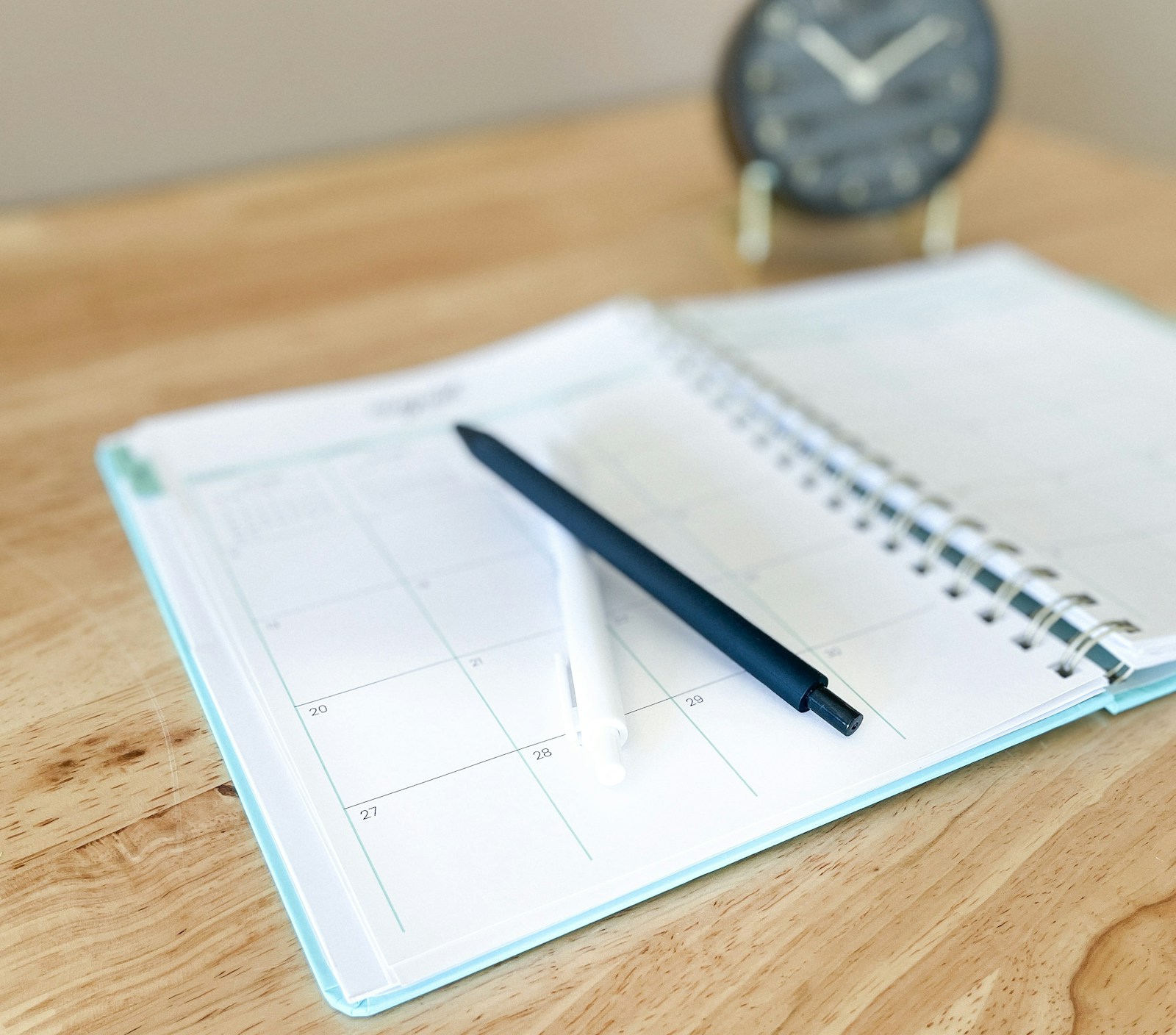 An open monthly planner with a black pen and a white pen on a wooden desk, with a small clock behind it
