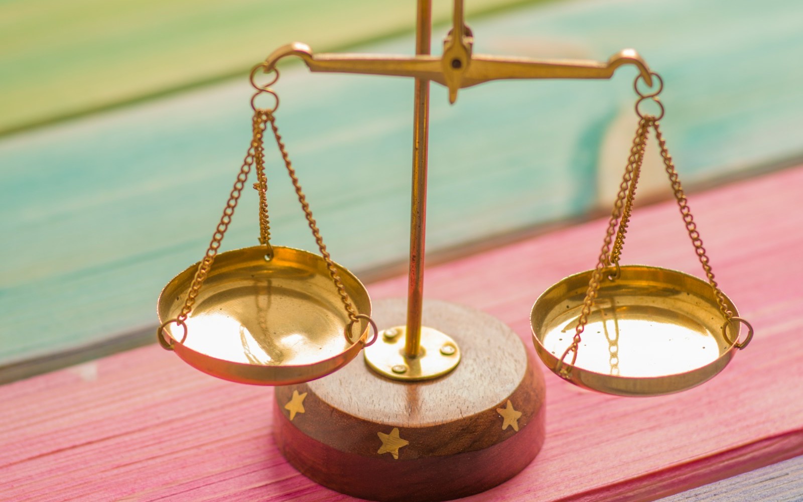 A small brass balance scale with two empty pans stands on a wooden base on pink and teal painted planks