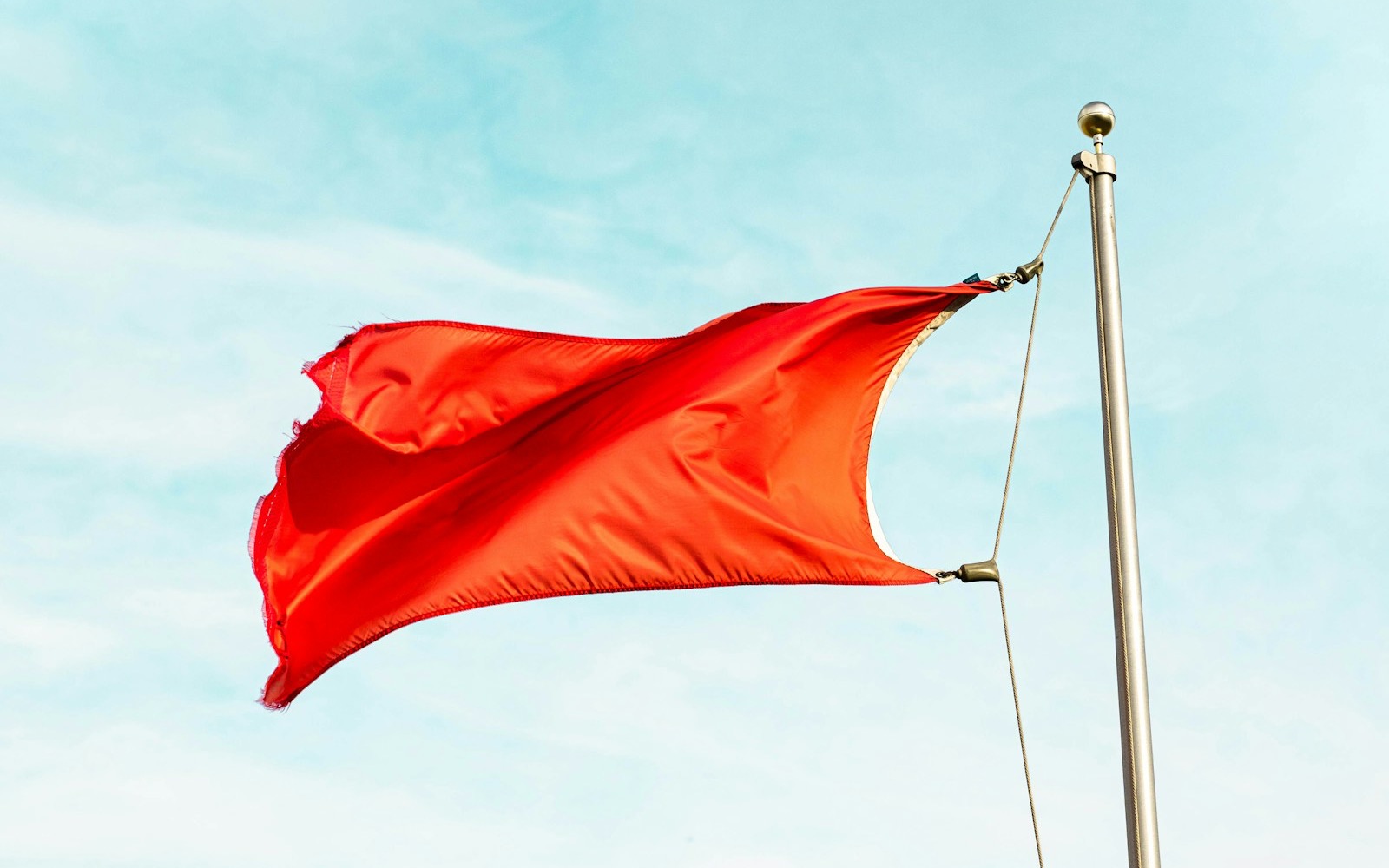 A red flag streams in the wind from a metal flagpole against a pale blue sky