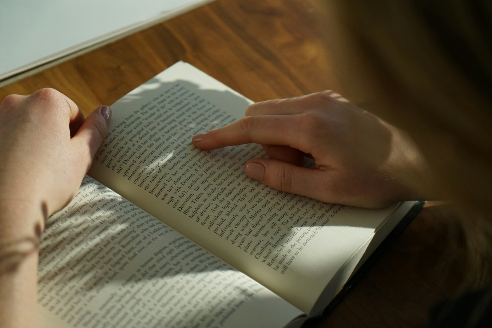 A person traces a line of printed text with one finger in an open book resting on a sunlit wooden table