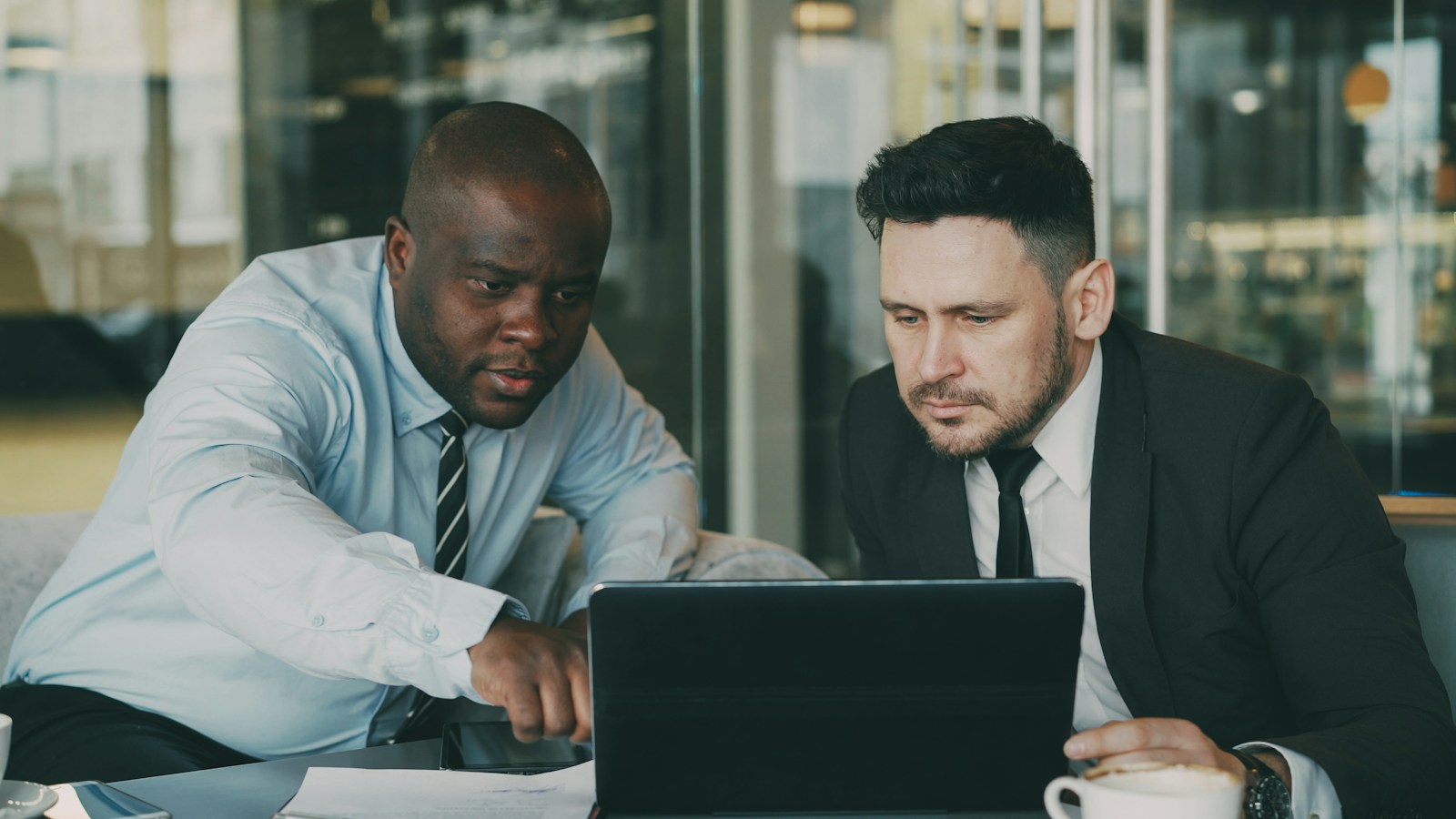 Two men in shirts and ties study a laptop screen at a cafe table while one of them points at it