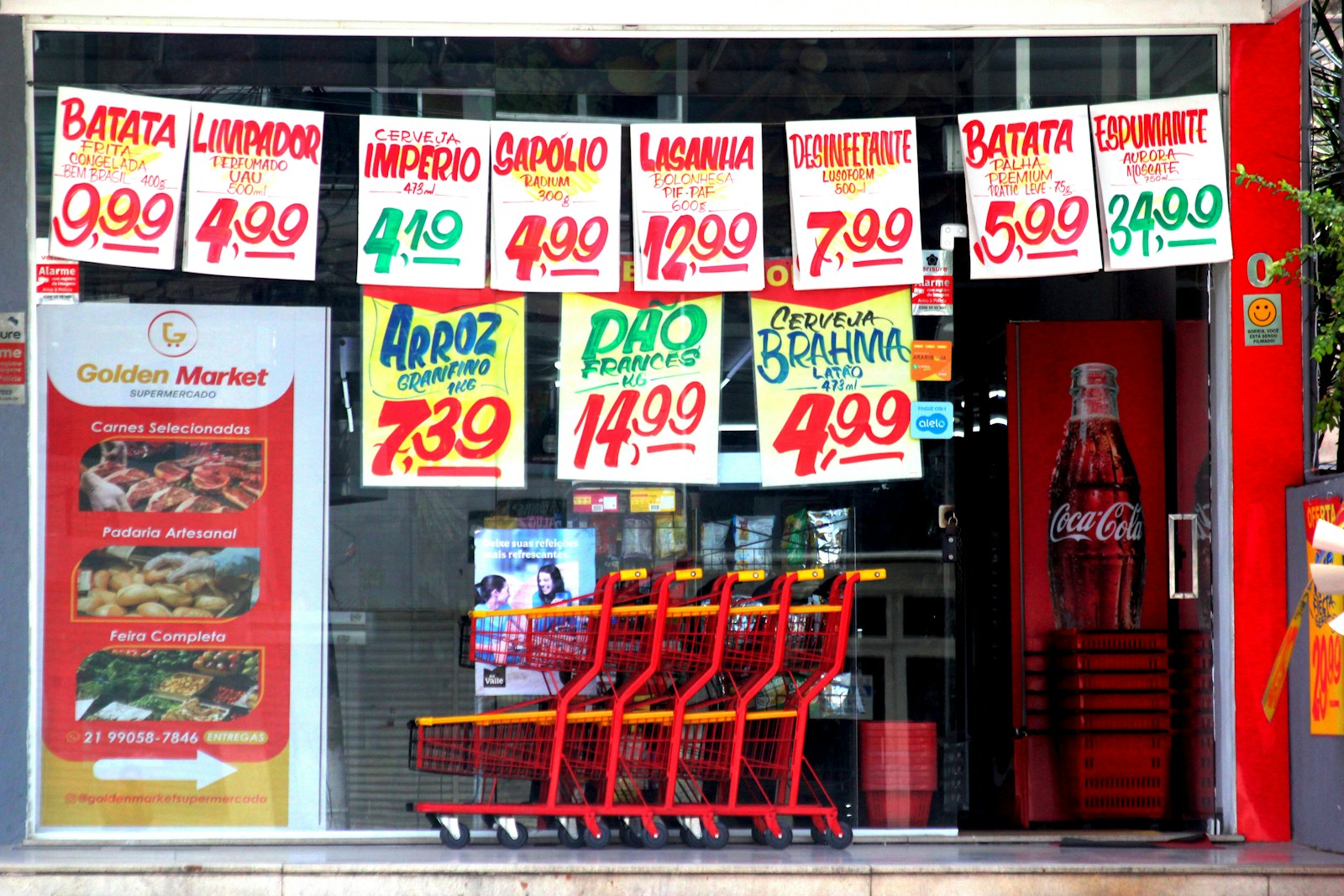 Hand-painted price signs for groceries hang in a shop window above a row of red shopping carts