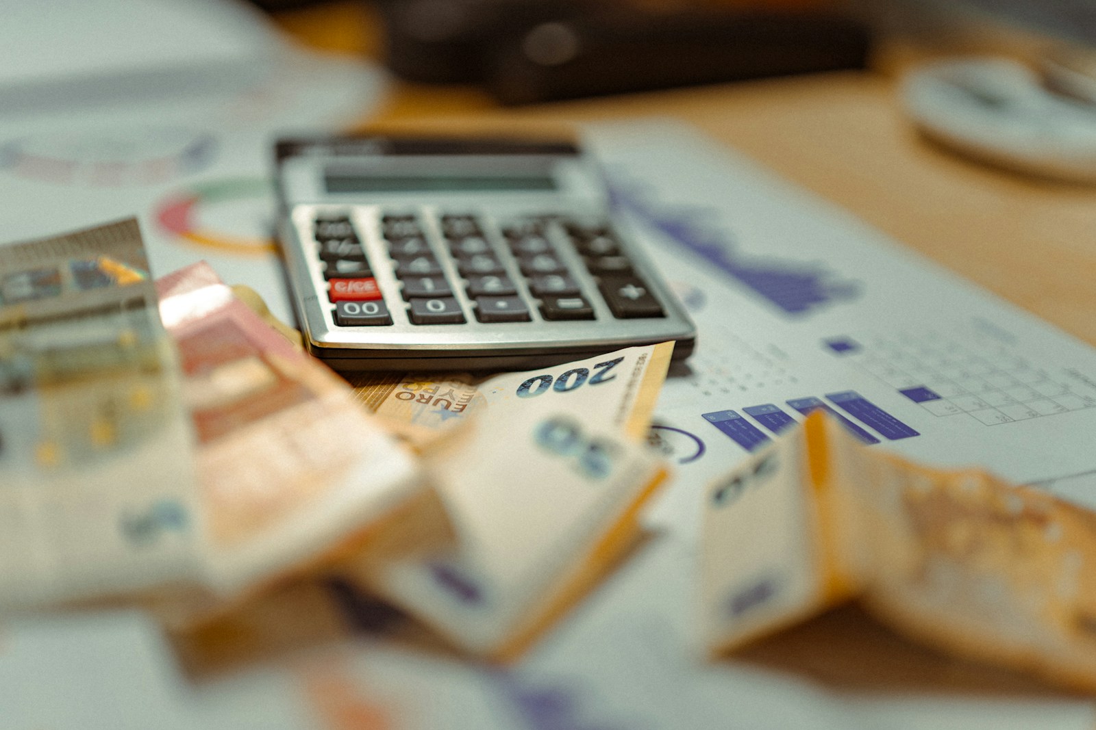 A silver desk calculator rests on printed bar charts beside folded euro banknotes on a wooden desk