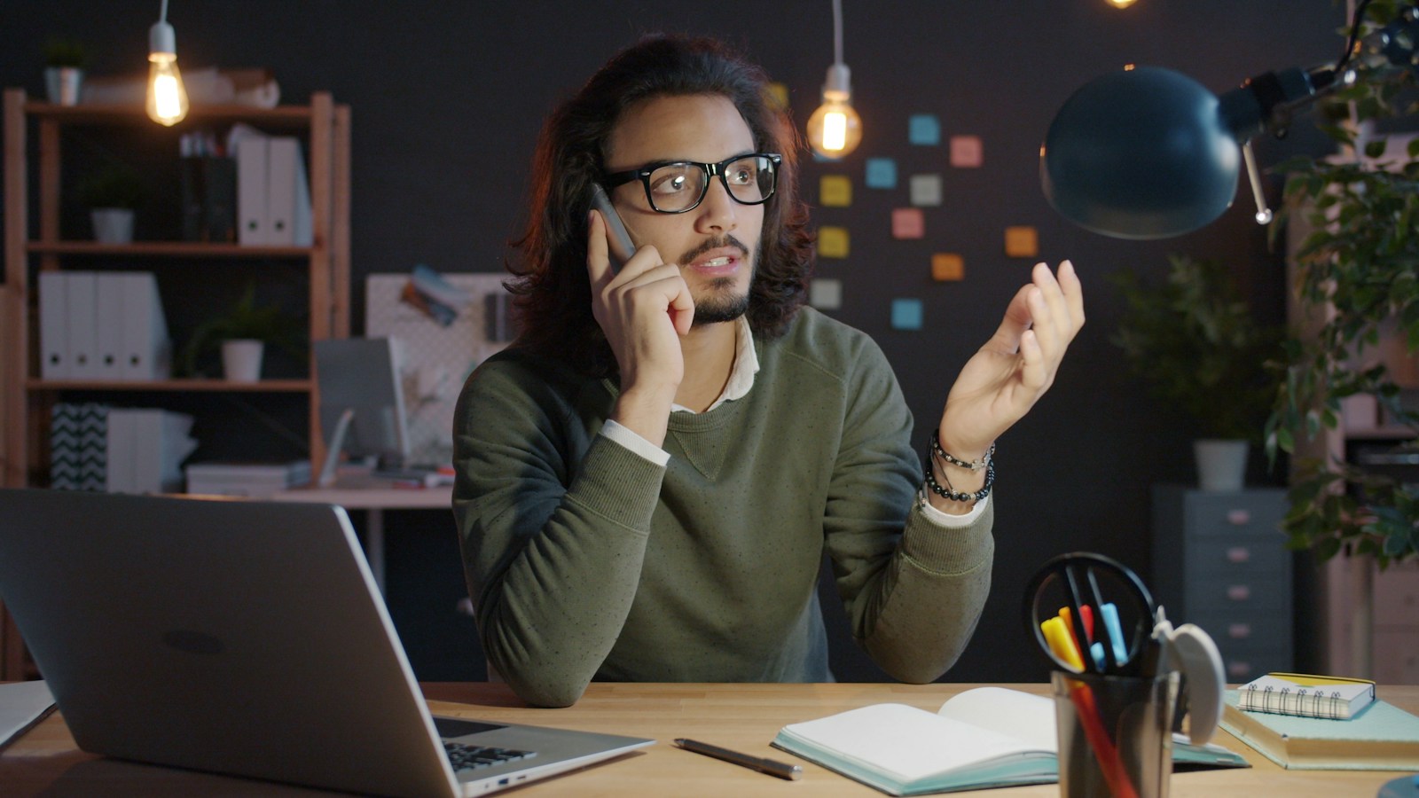 A man in glasses and a green sweater talking on a mobile phone at a desk with a laptop, notebook and desk lamp in an evening office