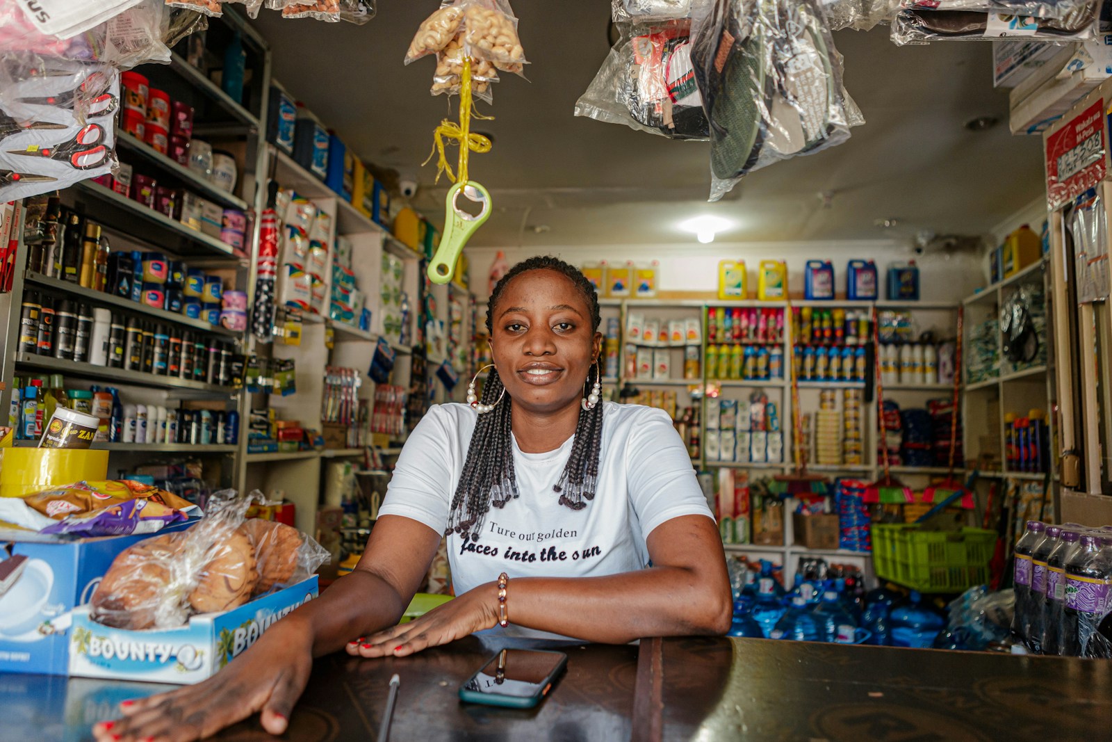 A shop owner in a white T-shirt leaning on her counter beside her phone, with shelves of groceries behind her