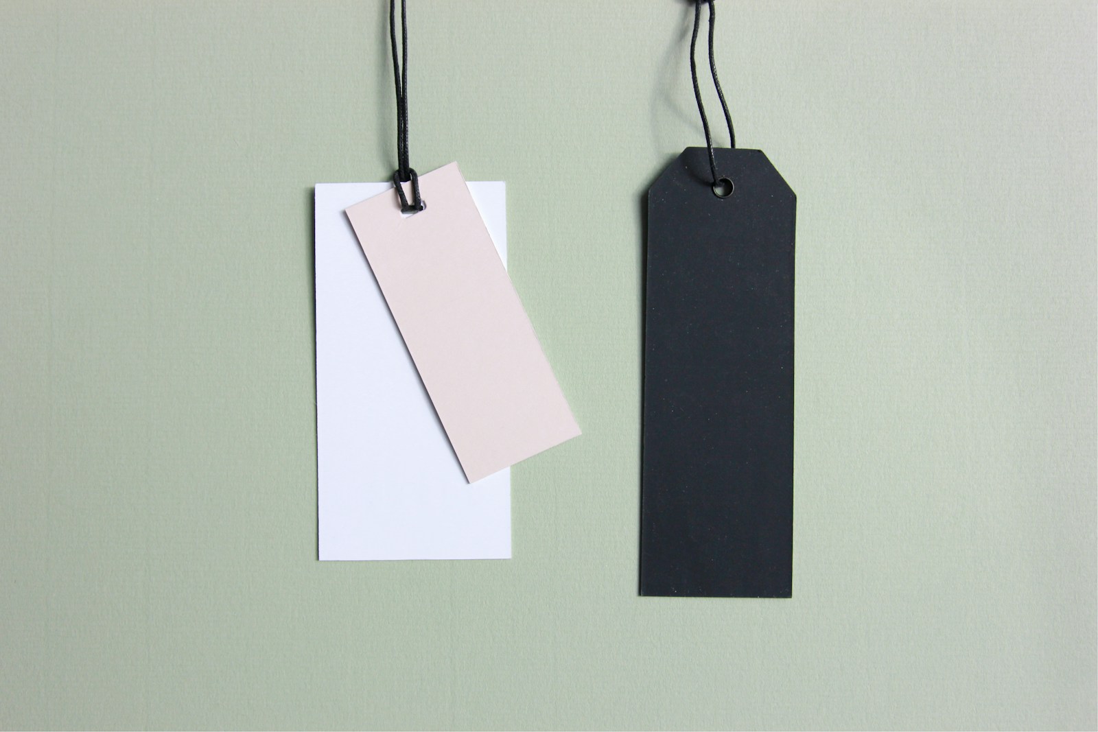Three blank paper tags, white, pale pink and black, hang on black strings against a sage green wall
