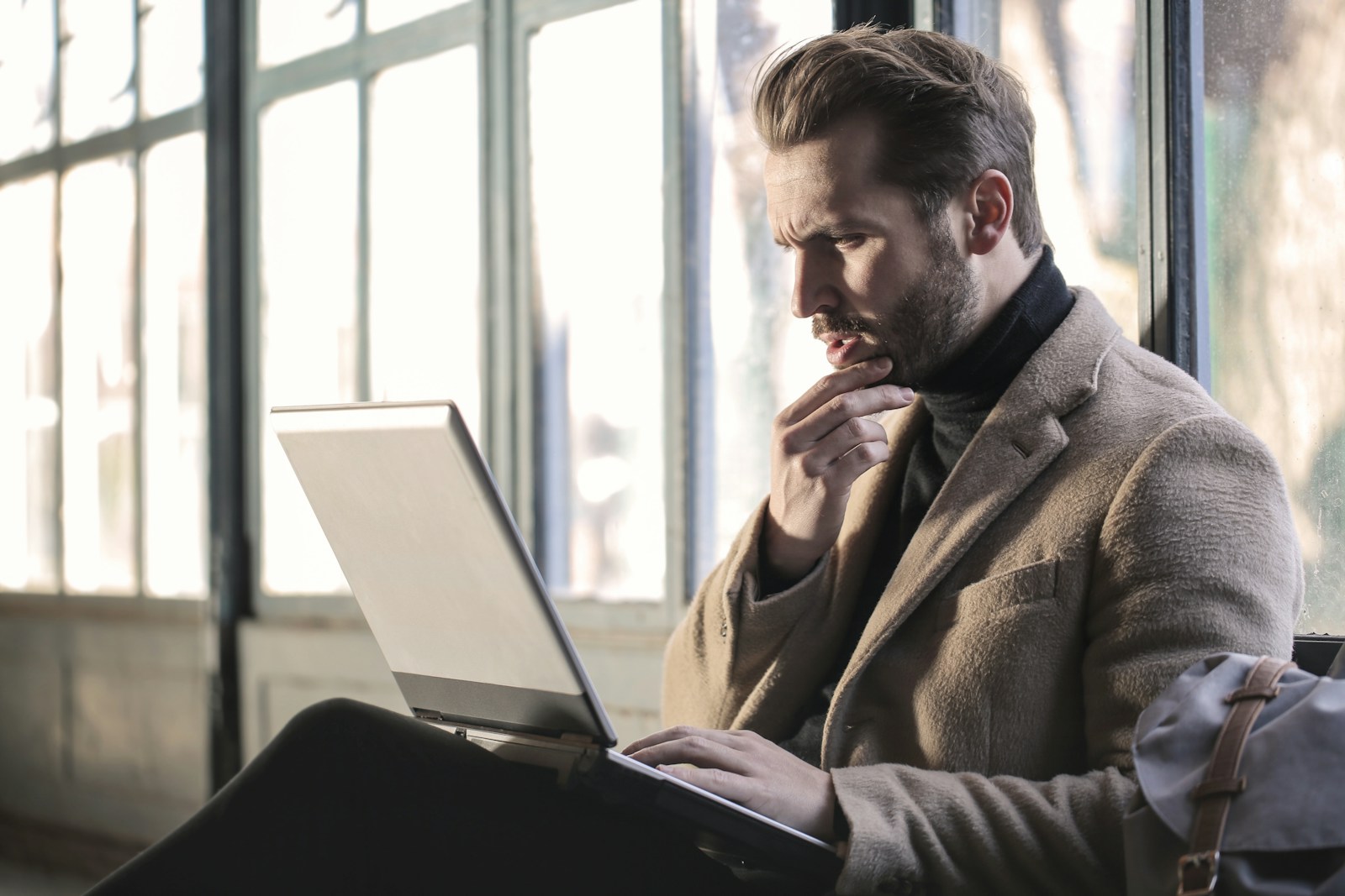 A bearded man in a camel coat frowns at his laptop screen with one hand on his chin by tall windows