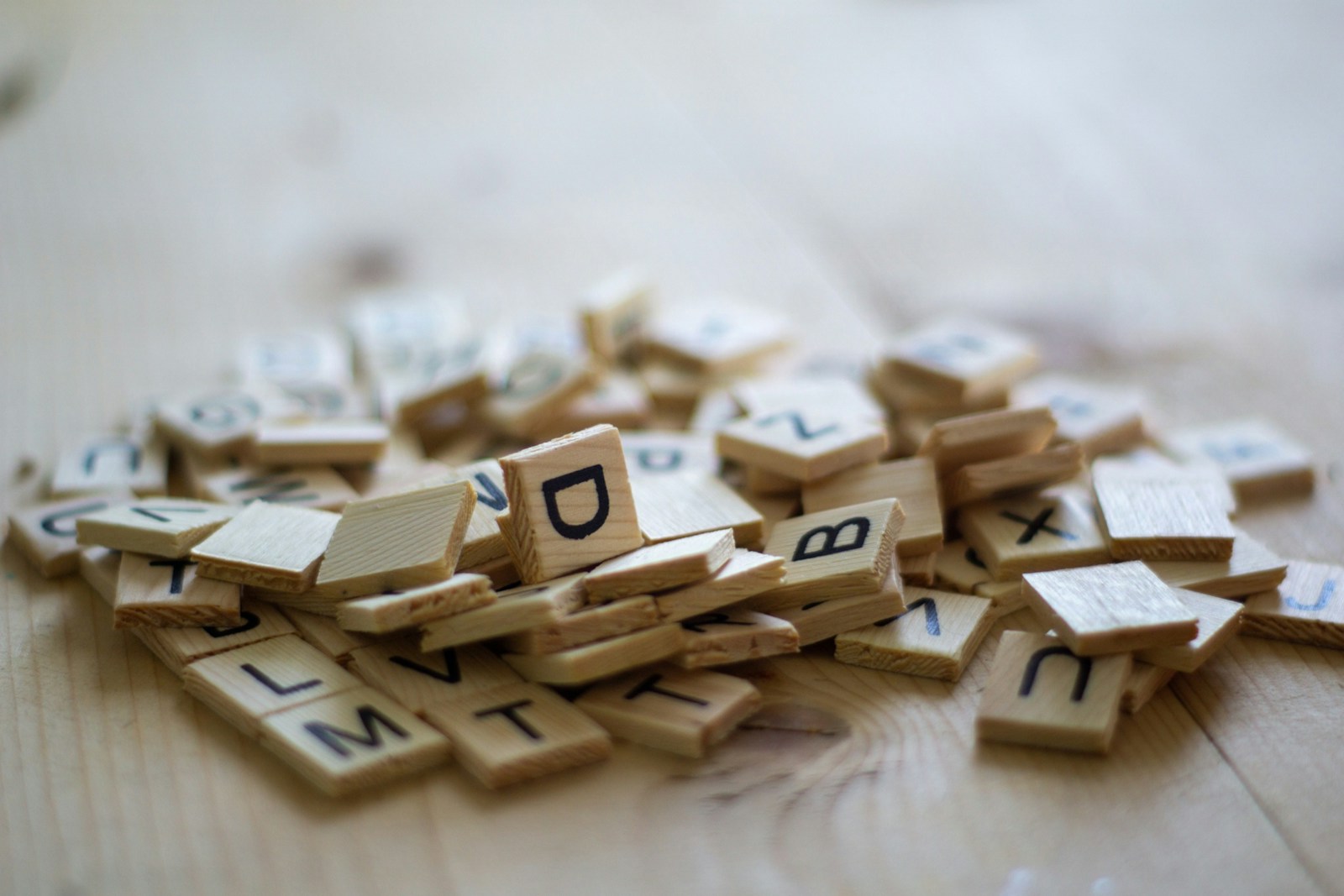 A pile of wooden letter tiles on a light wooden table, with the letters D, B, M and T facing up