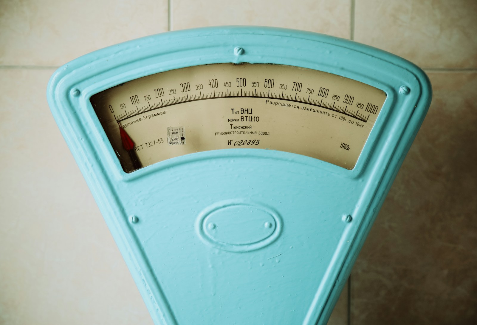 A vintage turquoise weighing scale with a dial marked from 0 to 1,000 against a tiled wall