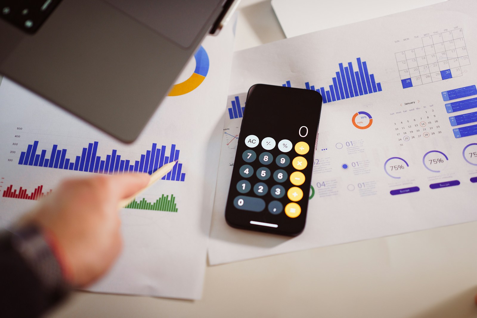 A hand holding a pencil points at printed bar charts next to a smartphone showing a calculator app