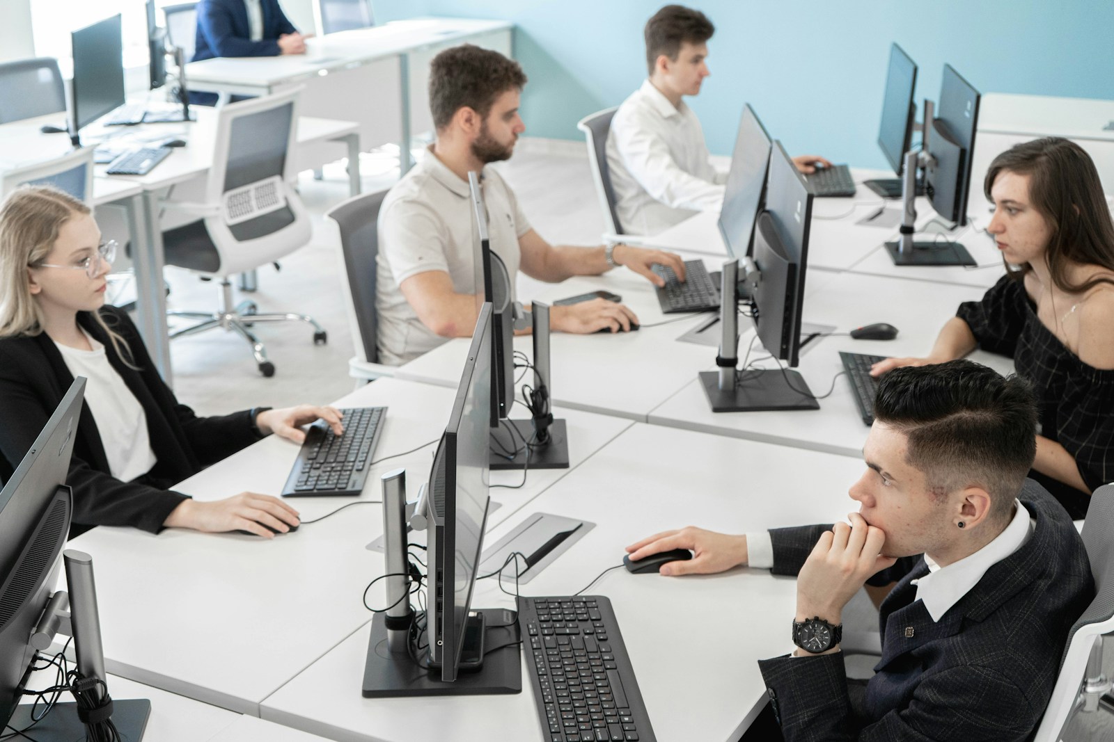 A team works at desktop computers on white desks in a bright office with a pale blue wall