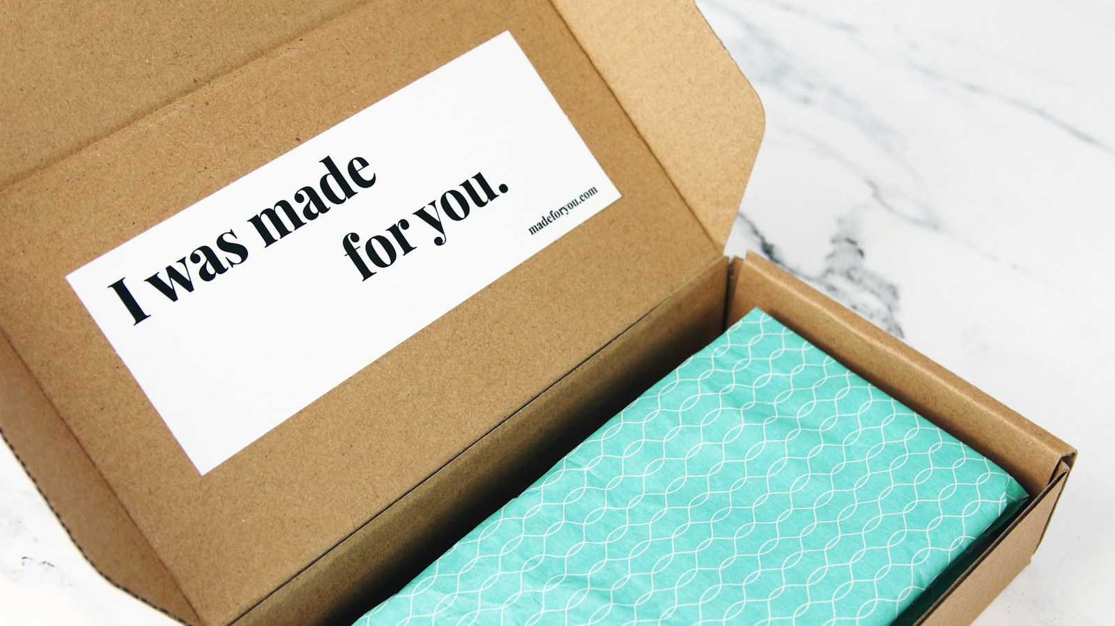 An open kraft mailer box on a marble surface with a printed thank-you card inside the lid and a product wrapped in teal tissue paper