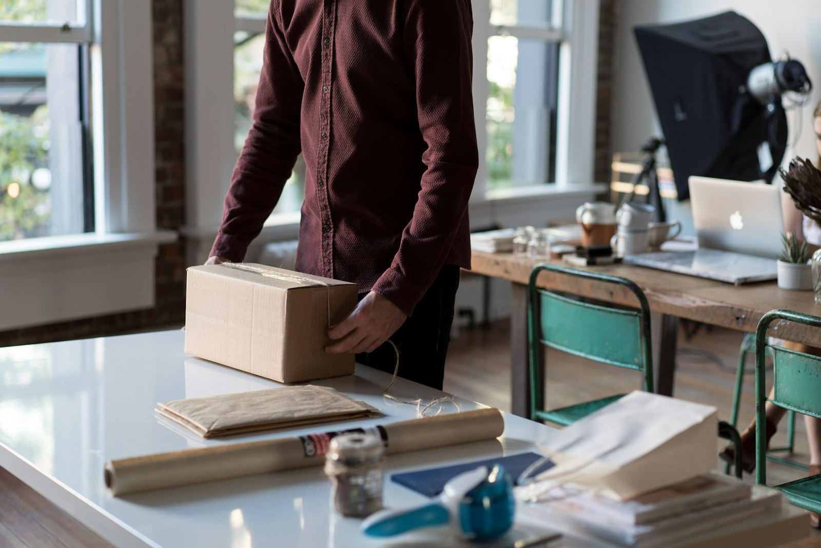 A man in a maroon shirt ties twine around a cardboard parcel on a white table in a bright studio, with kraft paper and a laptop behind him