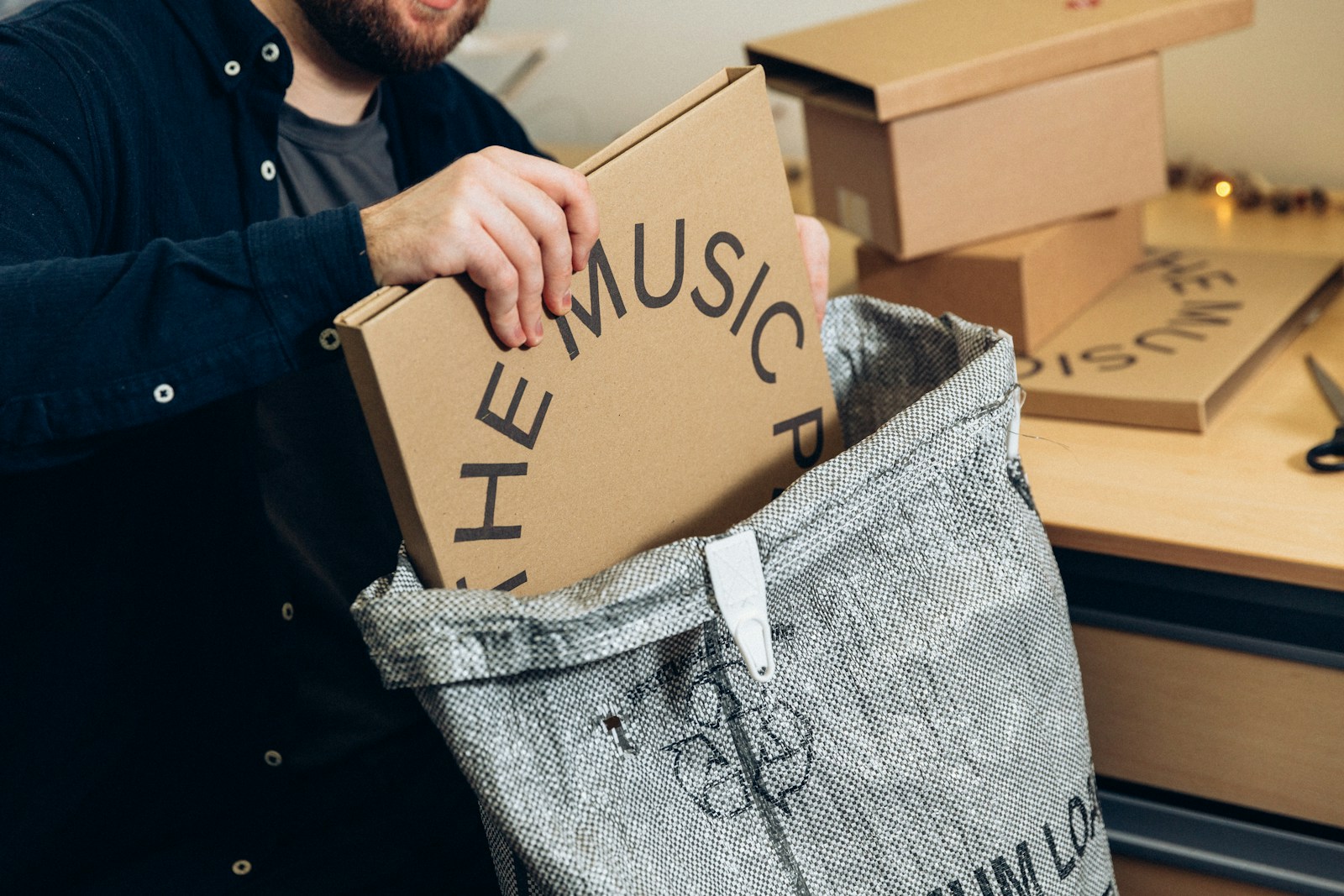 A bearded man slides a printed cardboard mailer box into a grey woven sack beside a desk stacked with more boxes