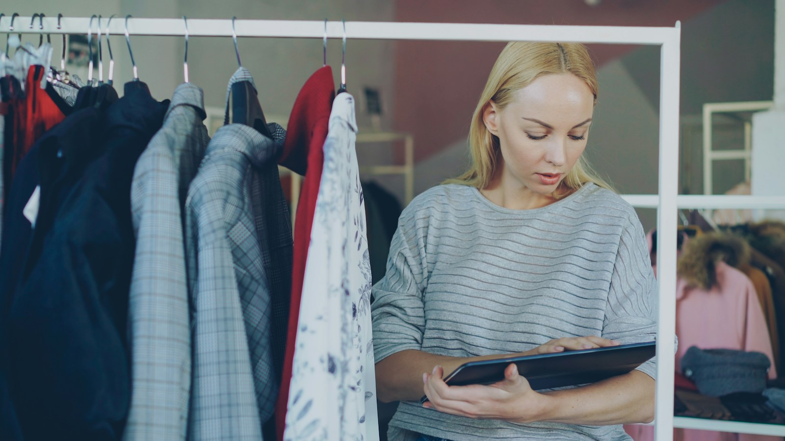 A woman in a grey striped sweater taps a tablet beside a rail of jackets in a clothing store