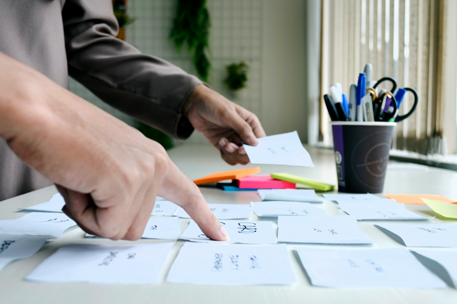 Hands arrange handwritten paper cards on a white desk, one finger pointing at a card, with sticky notes and a cup of pens behind