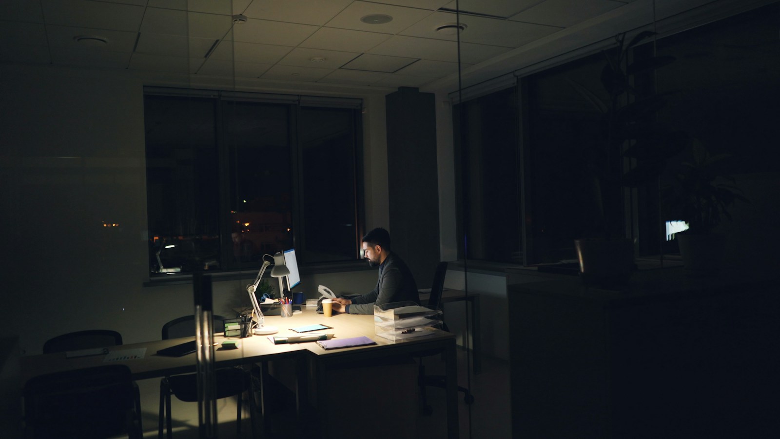 A man works alone at a lamp-lit desk in a dark office at night, with a coffee cup beside his monitor