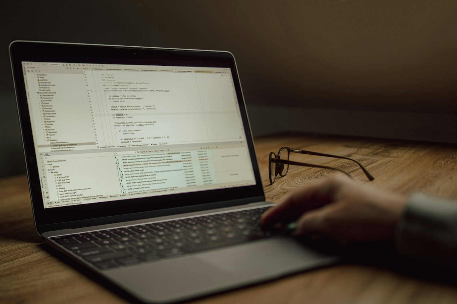 A hand on the keyboard of a laptop showing a code editor, with a pair of glasses on a wooden desk in a dim room
