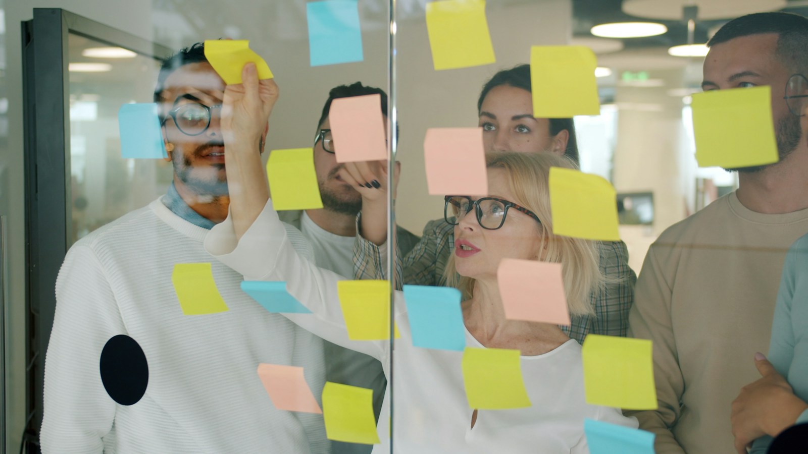 A team stands behind a glass wall covered in colourful sticky notes while a woman in glasses reaches up to place another note