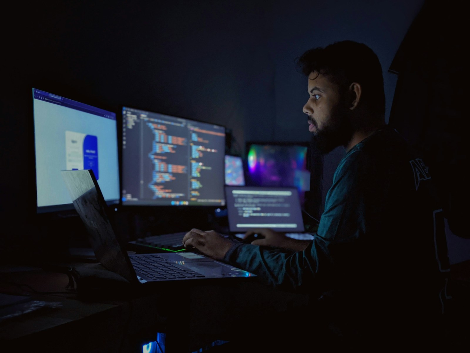 A bearded developer works on a laptop in a dark room, facing monitors full of code and a web page