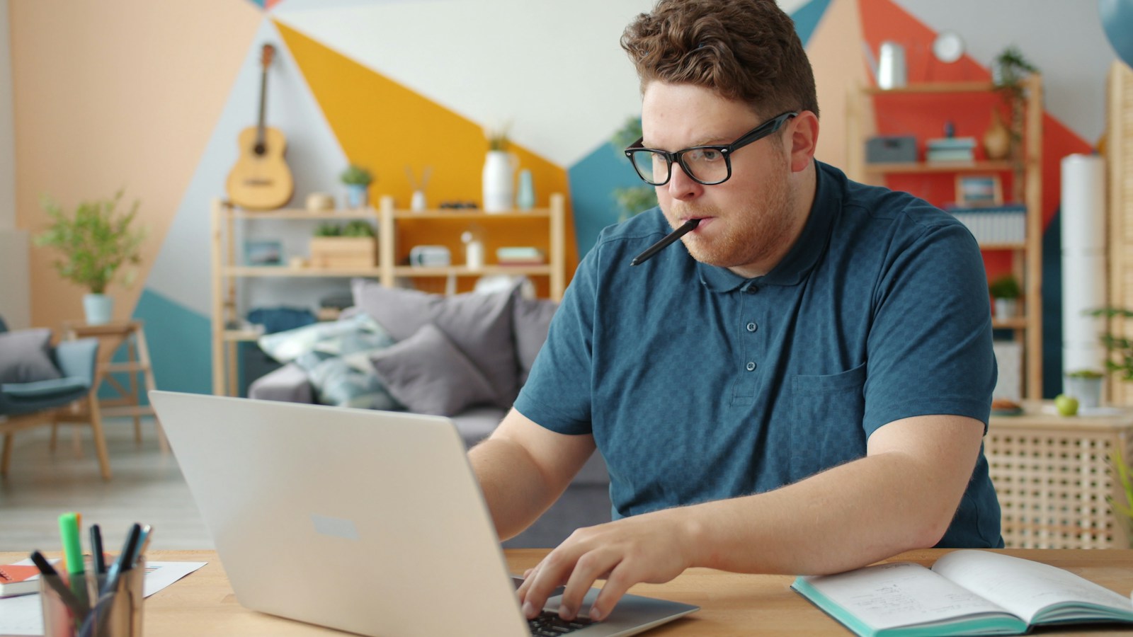 A man in glasses with a pen between his teeth types on a laptop beside an open notebook at a home desk
