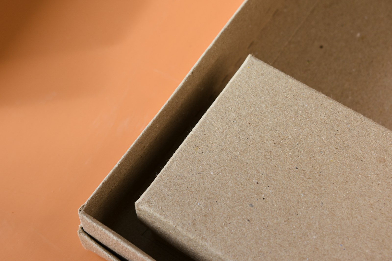 An open kraft cardboard box with a smaller kraft box resting inside it, photographed close up on an orange surface