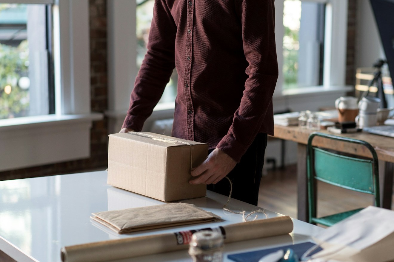 A man in a burgundy shirt holds a twine-tied cardboard parcel on a white packing table