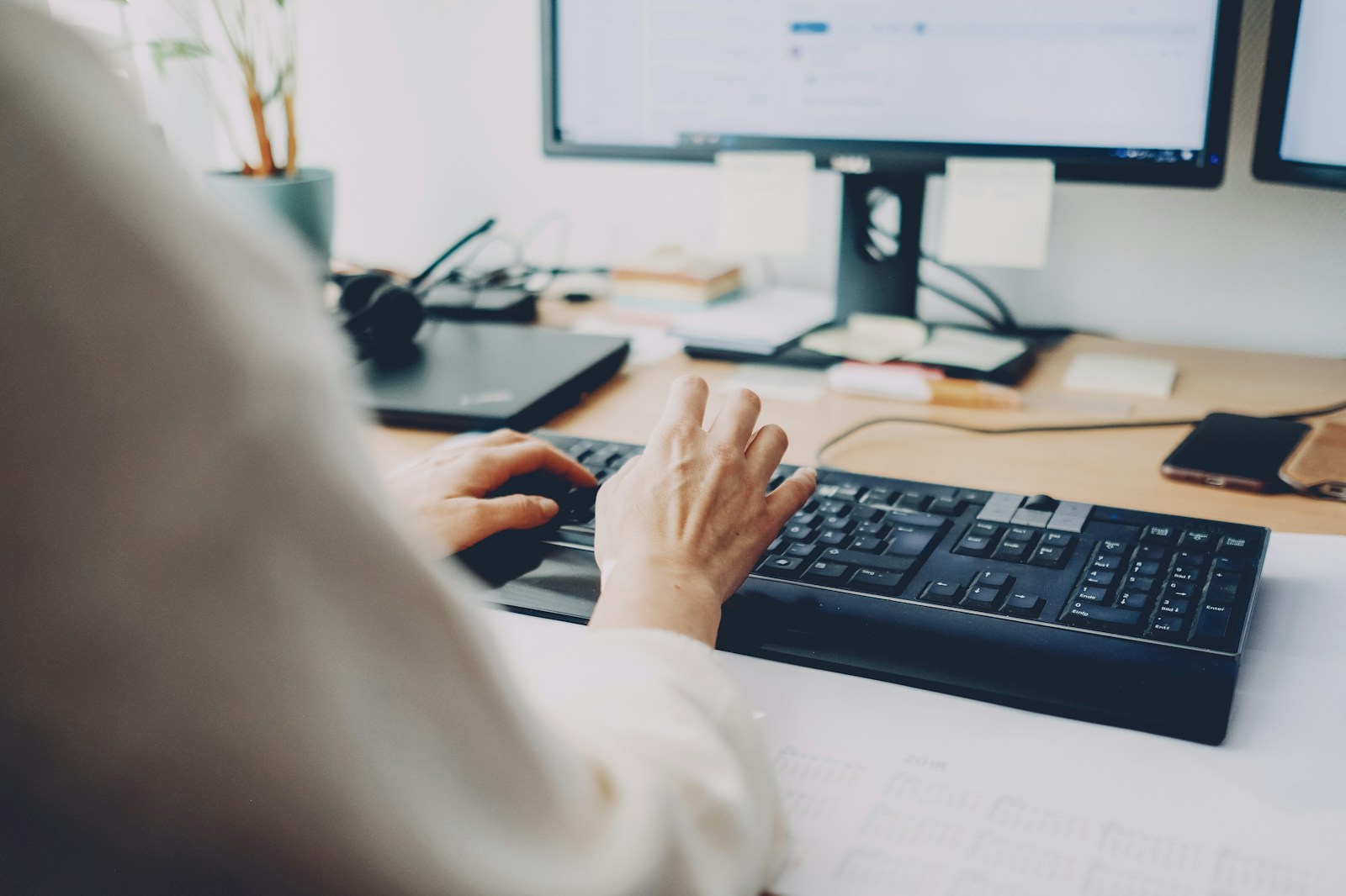 Over-the-shoulder view of a person typing on a black keyboard in front of a computer monitor on a wooden desk
