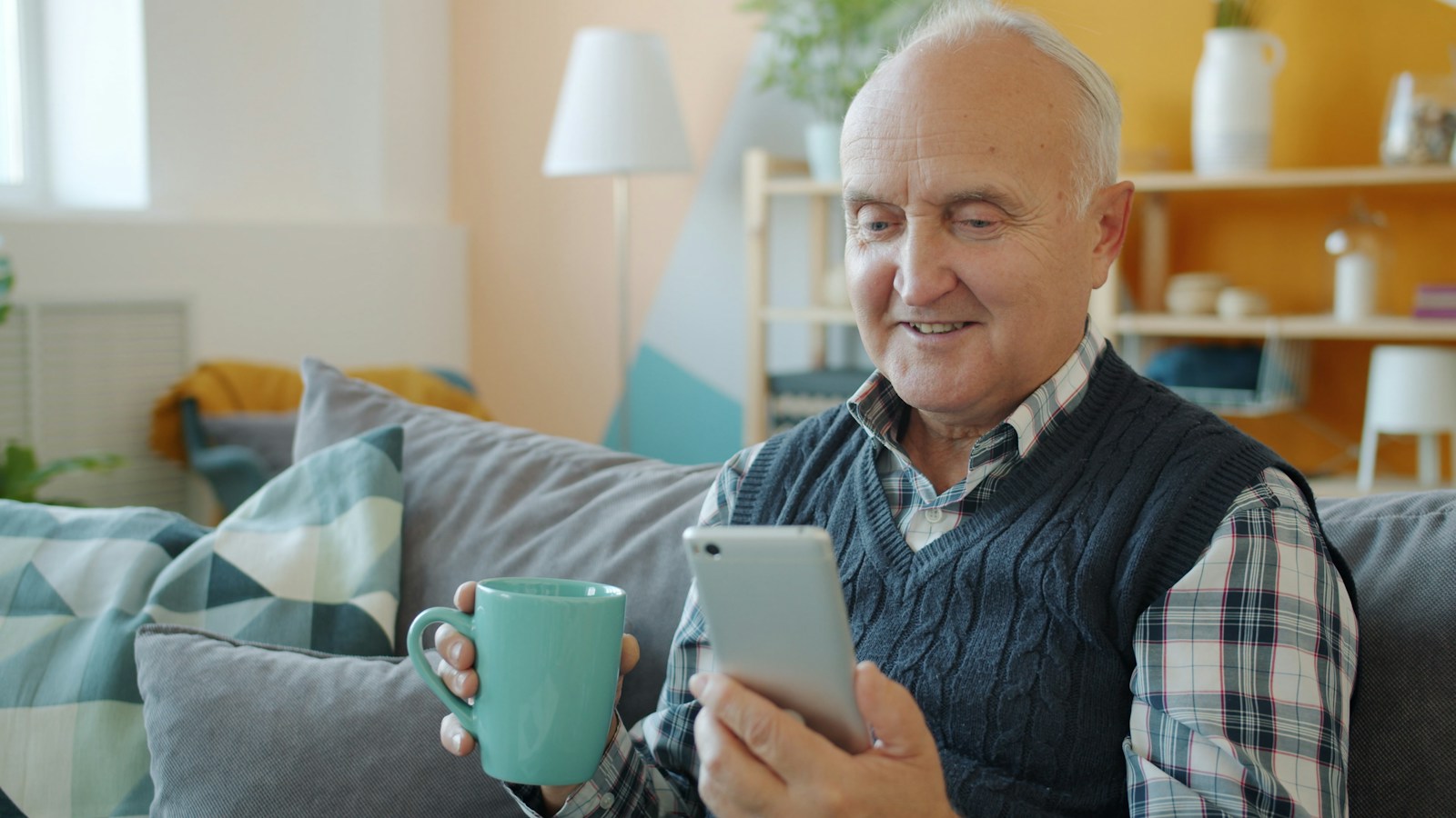 A smiling older man in a checked shirt and knitted vest reads a smartphone on a sofa while holding a teal mug