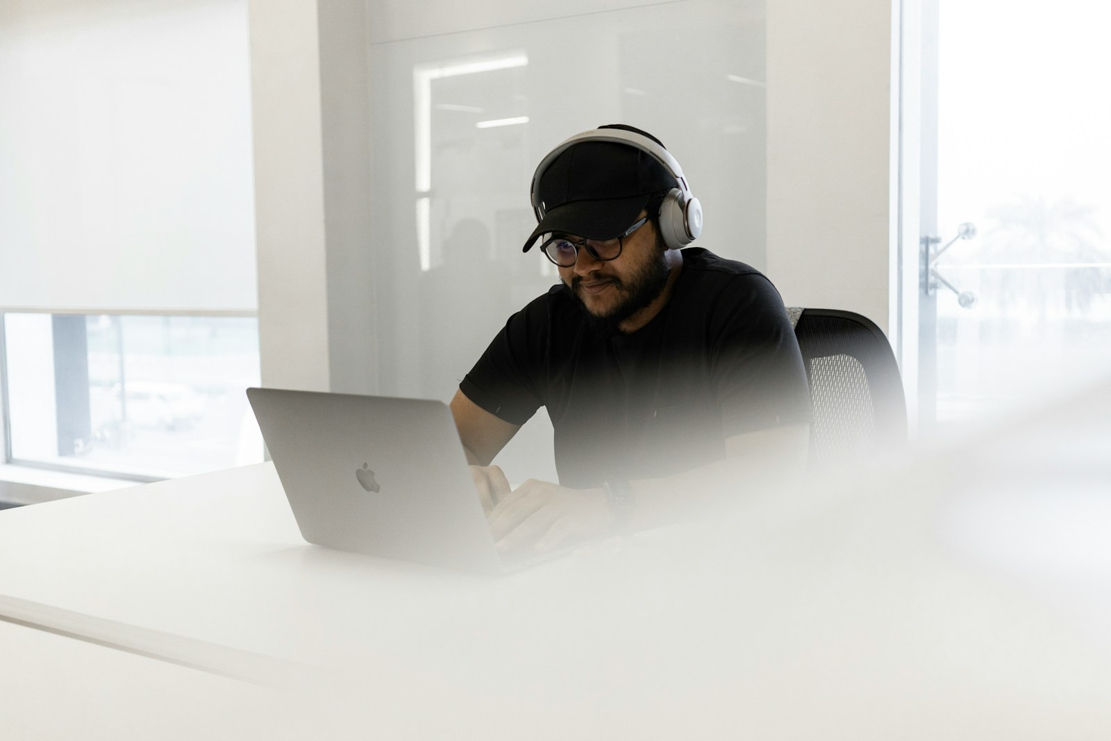 A man in a black cap, glasses and white over-ear headphones types on a laptop at a white desk