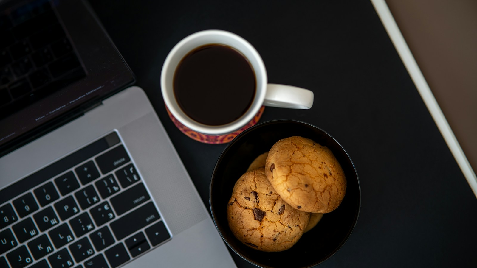 A laptop keyboard beside a cup of black coffee and a bowl of chocolate chip cookies on a dark desk