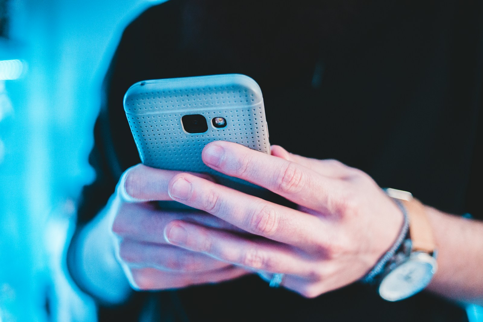 Two hands holding a grey smartphone in a perforated case under cool blue light while someone uses it