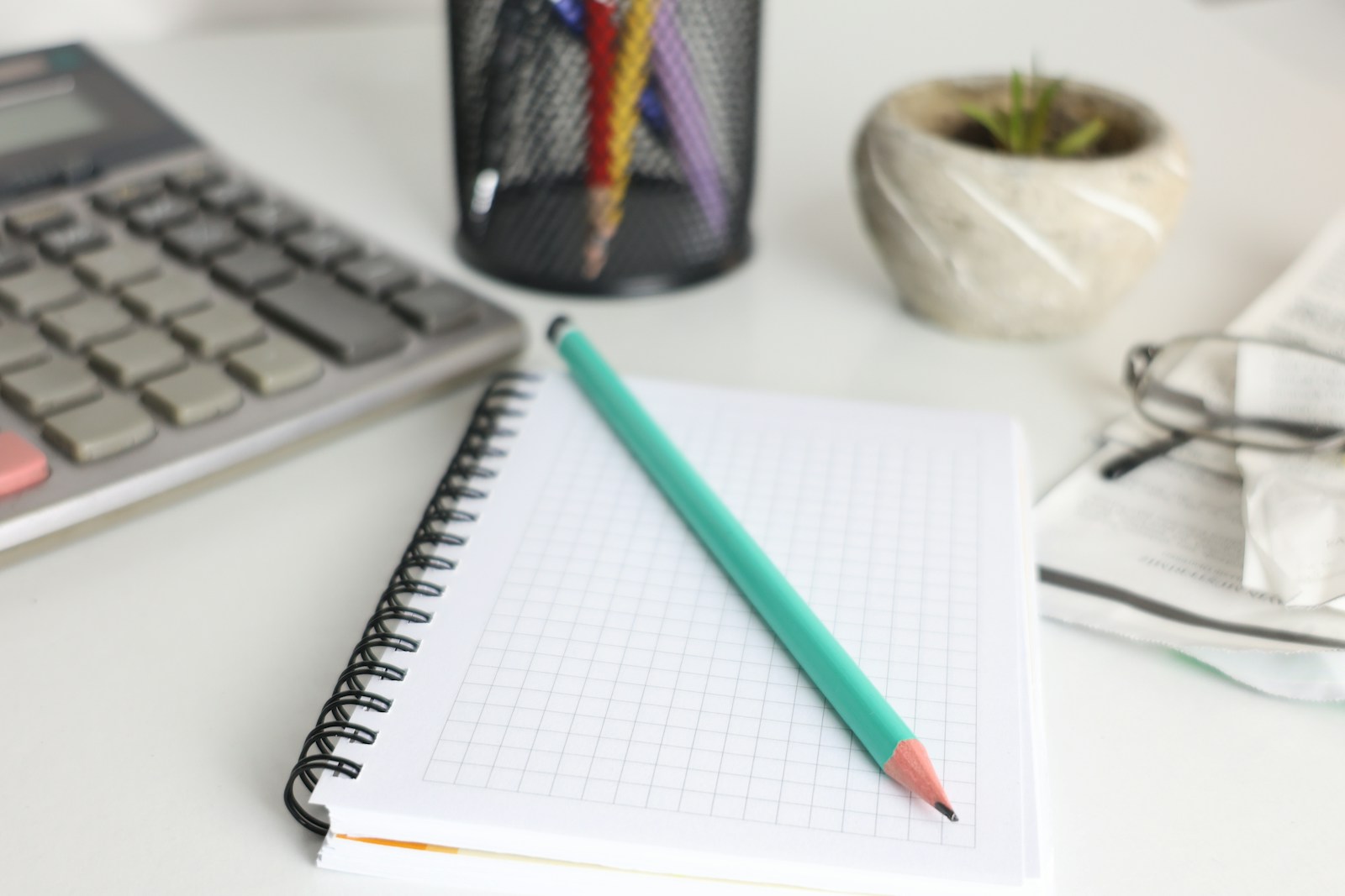 A calculator, a grid notebook with a green pencil, a pen pot and a small potted plant on a white desk